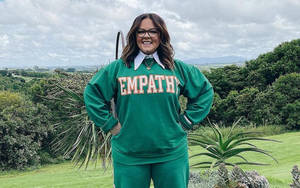 穿著綠色 'EMPATHY' 運動衫的女性在戶外長滿青草的山丘上微笑，背景有樹木和山脈。