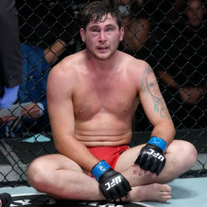 疲憊的UFC拳手在比賽後滿臉鮮血、手臂有紋身，坐在八角籠地板上。 — 運動 壁紙