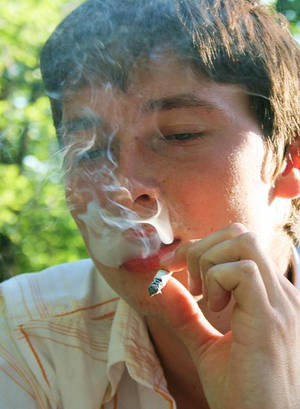 年輕男子在戶外明亮的陽光下吸煙，身後有綠色樹木。