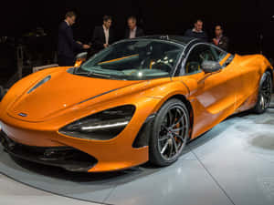 橙色McLaren 720S超級跑車在車展上展出，旁邊有身穿西裝的男士。 — 汽車 壁紙