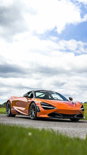 橙色McLaren 720S跑車在碎石路上，有草和多雲天空。