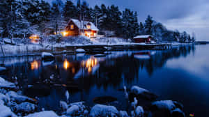 白雪覆蓋的湖邊小屋，溫暖的燈光在夜晚平靜的水面上反射。