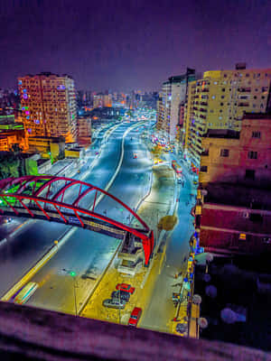 城市夜景，一座紅色橋樑橫跨繁忙的高速公路，建築物燈火通明。