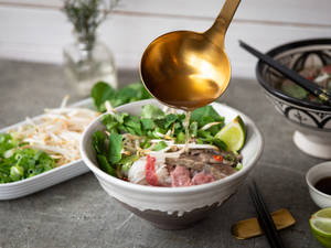 一碗冒著熱氣的越南河粉，湯汁倒在牛肉、香草和青檸上。