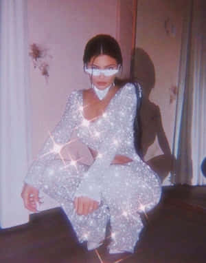 身穿閃亮銀色服裝的女性，戴著白色太陽眼鏡蹲在室內。