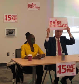 兩人在桌子旁舉著支持 raise chicago 和 15 cant wait 的抗議標語牌。 — 政治 壁紙