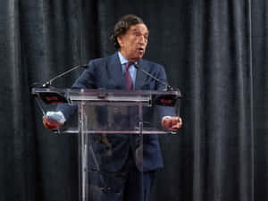 穿西裝的男子在講台發言，手持口罩。