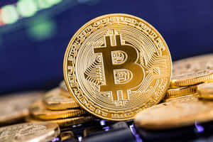 一枚帶有電路圖案的金色比特幣硬幣放在一堆類似硬幣上，靠近發光的屏幕。 — 比特幣 壁紙