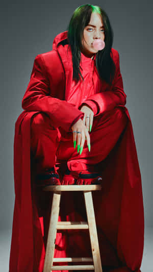 Billie Eilish 身穿紅色服裝在木凳上吹粉紅色泡泡糖泡泡。
