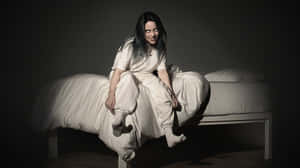 Billie Eilish 坐在黑暗房間的床上，身穿白色休閒服。