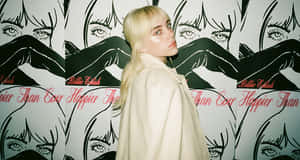Billie Eilish 身穿米色外套，在她的專輯藝術牆前擺姿勢。