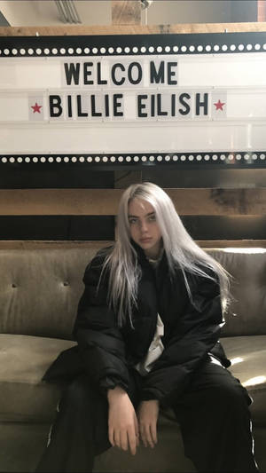 Billie Eilish 坐在梳化上，下方有一個帶有她名字的歡迎招牌。