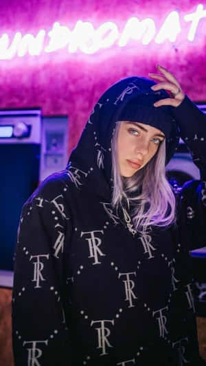 Billie Eilish 穿著帶有白色標誌的黑色連帽衫，在粉紅色霓虹燈下。