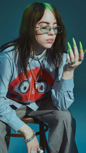 Billie Eilish 在攝影棚內戴著眼鏡和綠色指甲坐著。