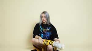 Billie Eilish 在攝影棚內盤腿而坐，身穿黑色圖案T恤和黃色褲子。