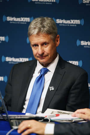 身穿西裝和藍色領帶的男子在SiriusXM活動中，面前有咪高峰和文件。
