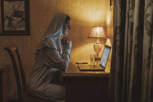 戴頭巾的女性在溫暖燈光下，在桌上與筆記本電腦一起祈禱。