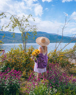 戴草帽的女性在湖邊手持鮮花，背景有山脈