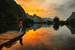 夕陽下，竹筏上站立的人，平靜水面倒映著山巒。
