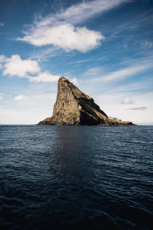 一座陡峭的岩石島嶼獨自矗立在開闊的海洋中，天空部分多雲。