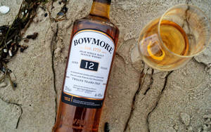 Bowmore威士忌瓶和玻璃杯在沙灘上，旁邊有海藻。