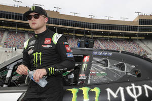身穿黑色賽車服的NASCAR車手，在體育館內手持水樽站在賽車旁。