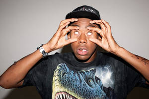 年輕男子戴著帽子和恐龍T恤，用手俏皮地框住眼睛。 — Tyler the creator GIF 壁紙
