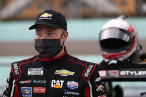 NASCAR車手身穿賽車裝備，頭盔放在身旁，在日落時分的賽車場上。