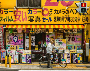騎單車的男人在色彩繽紛的日本相機店前停下來，店內有促銷標誌。 — 藝術 壁紙