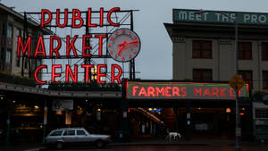 Public Market Center和Farmers Market的霓虹燈招牌在黃昏時發光，附近有一輛銀色汽車和一隻狗。