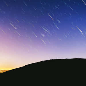 星軌在暮色天空中劃出弧線，在黑暗山丘之上，城市燈光在地平線閃爍。 — 天空美學 壁紙