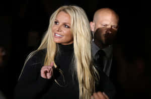 Britney Spears 穿著黑色高領毛衣微笑，身後有穿西裝的男子。