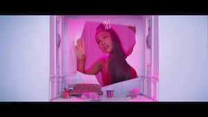 Ariana Grande 喺一個鋪滿白色絨毛地板、粉紅色燈光嘅娃娃屋房間入面。