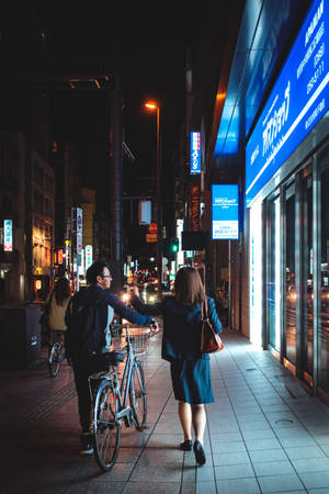 情侶在夜晚沿著霓虹燈閃爍的城市街道行走，帶著一輛單車