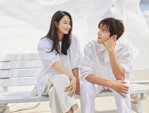 一對微笑的情侶穿著白色衣服，坐在海灘上白色天篷下的長椅上。 — 你的名字 壁紙