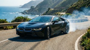 黑色BMW i8在海邊道路上飄移，背景有海洋和綠色山丘。