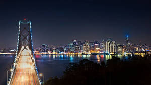 夜間城市景觀，包括燈火通明的海灣大橋和舊金山天際線，水面倒影。 — 旅行 壁紙