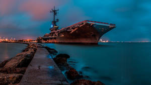 一艘大型航空母艦在黃昏時停泊，背景有平靜的水面和城市燈光。 — 海洋 壁紙