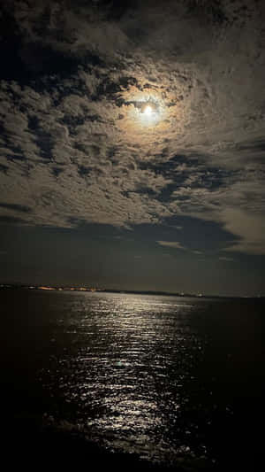 月光在水面上反射，多雲的夜空下遠處有城市燈光。 — 夜空 壁紙