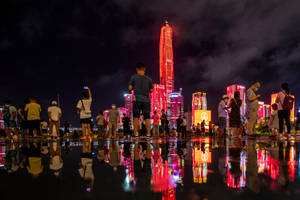 夜間城市景觀，人們觀看一座發光的紅色摩天大樓反射在潮濕的人行道上。 — 唐人街 GIF 壁紙