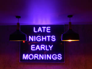 紫色霓虹燈牌，上面寫著「late nights early mornings」，位於兩盞吊燈之間。