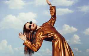 身穿金色外套和太陽眼鏡的女性，背後有雲朵，伸手觸及天空。