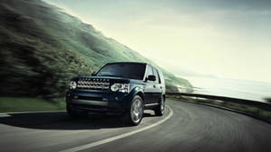 黑色 Land Rover SUV 在海岸道路上行駛，背景有模糊的綠色山丘和海洋