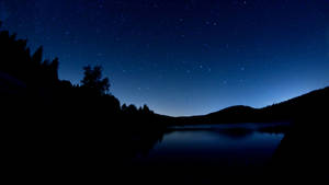 平靜湖面倒映星空夜空，伴隨黑暗樹木和山脈剪影。 — 深藍色 壁紙