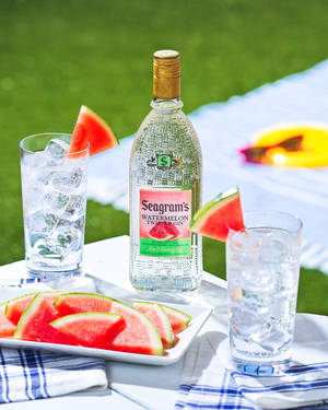 Seagram's Watermelon Gin 瓶子連同杯子和西瓜片放在戶外野餐桌上。 — 電腦 GIF 壁紙