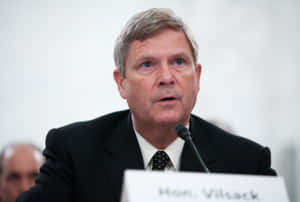 身穿西裝的男子在寫有 Hon Vilsack 的講台上發言 — 政治 壁紙