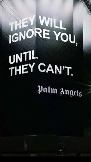 深色牆上以粗體白色文字寫著「THEY WILL IGNORE YOU UNTIL THEY CAN'T」及下方「Palm Angels」。