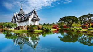 白色泰式寺廟倒映在平靜的池塘中，周圍有綠樹和藍天
