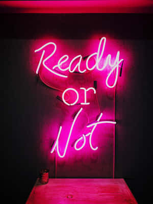 粉紅色霓虹燈牌，上面寫著「Ready or Not」，掛在黑暗的牆壁上，下面有一張木桌。
