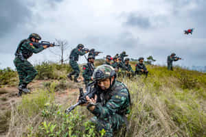 身穿迷彩裝備的士兵在陰天天空下穿越草坡，一架無人機在頭頂飛過。 — 戰爭 壁紙